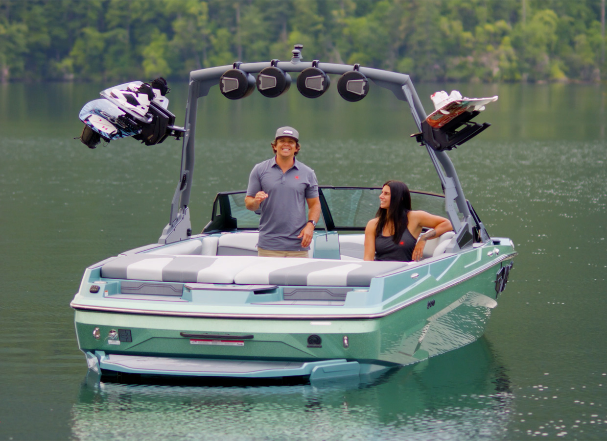 Two people stand smiling in the cockpit of a light teal Axis A200 floating on a calm lake, wakeboards on the tower rack and forested hills behind.