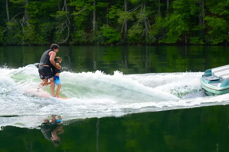 An adult wakesurfs while holding a small child on the board behind a seafoam-green Axis A200 on a calm, tree-lined lake.