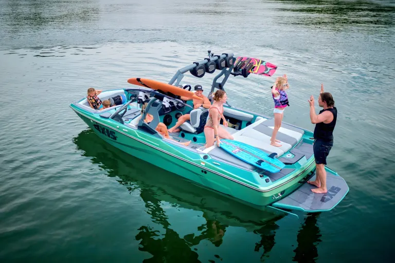 A seafoam-green Axis A200 idles on a calm forested lake, friends aboard and one standing on the rear platform, boards racked on the tower.