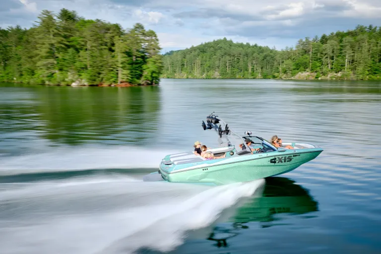 A seafoam-green Axis A200 runs across a glassy lake throwing spray, passengers aboard and a forested shoreline under a cloudy sky.