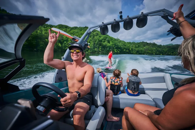A smiling driver points skyward from the helm of an Axis A200 as children watch a wakesurfer behind, gray-and-seafoam interior on a forested lake.