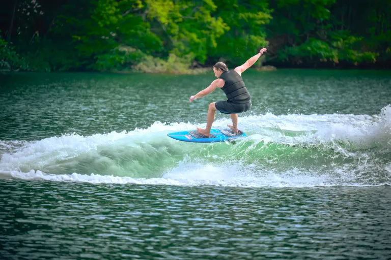 A wakesurfer in a black vest carves the clean green wake behind an Axis A200 on a forested lake.