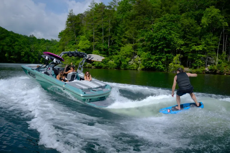 A seafoam-green Axis A200 tows a wake foiler riding a blue board across a tree-lined lake, passengers watching from the cockpit.