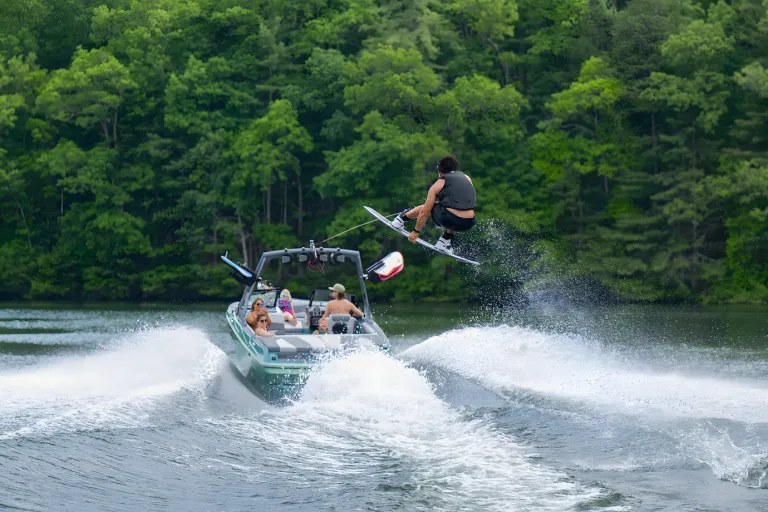 A wakeboarder boosts high off the wake behind a seafoam-green Axis A200, spray flying against a forested shoreline.