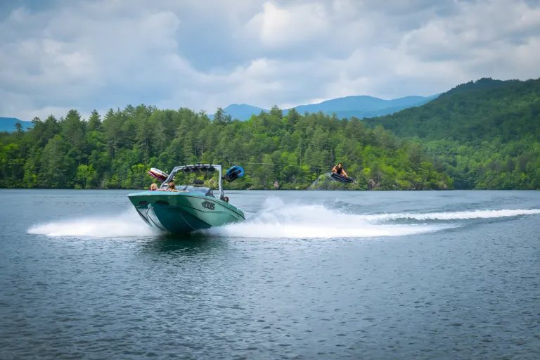A seafoam-green Axis A200 runs across a wide mountain lake with a wakesurfer riding its wake in the distance.