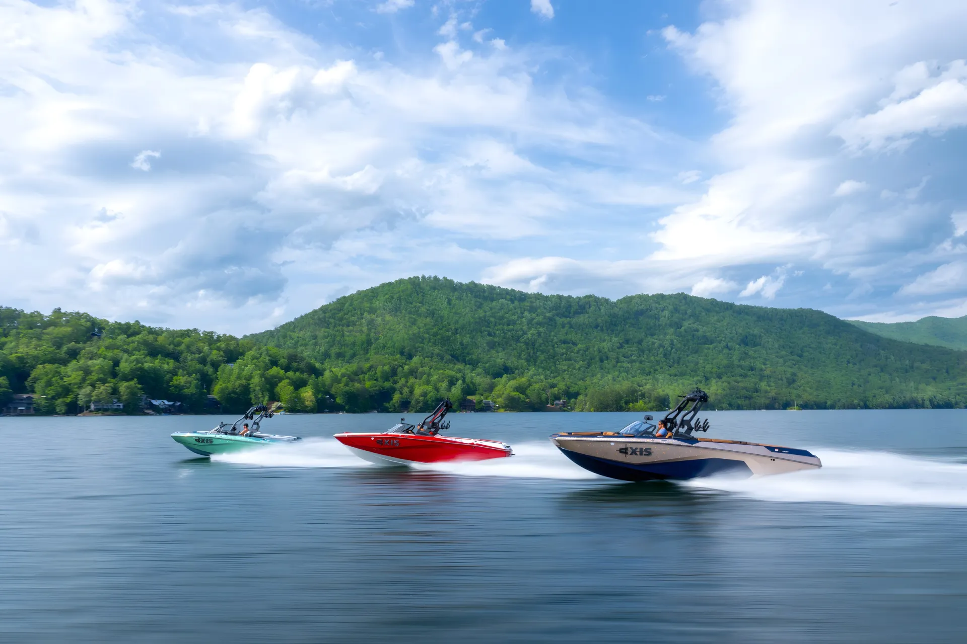 Three Axis A-Series wake boats — seafoam, red and black — running in formation across a lake below a forested hill.