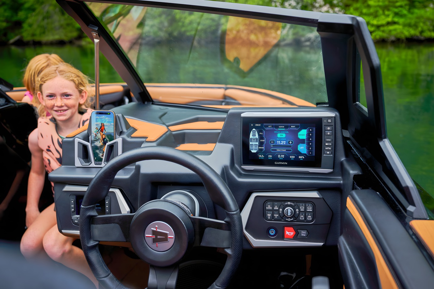 A red-haired girl kneels smiling beside the Axis helm, the Garmin touchscreen and Axis-logo wheel in view, tan interior and a tree-lined green cove behind.