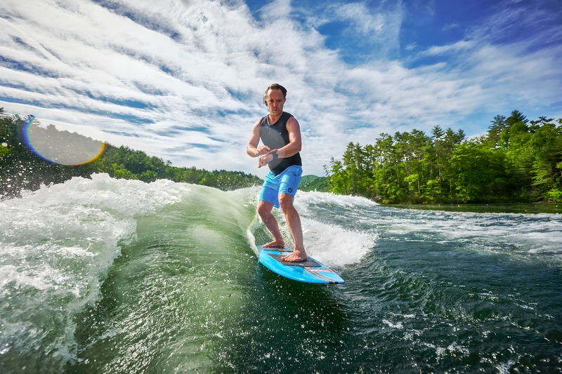 A man wakesurfs a clean green wave on a blue board under a bright sky, seen from the back of the boat with a forested shoreline behind.