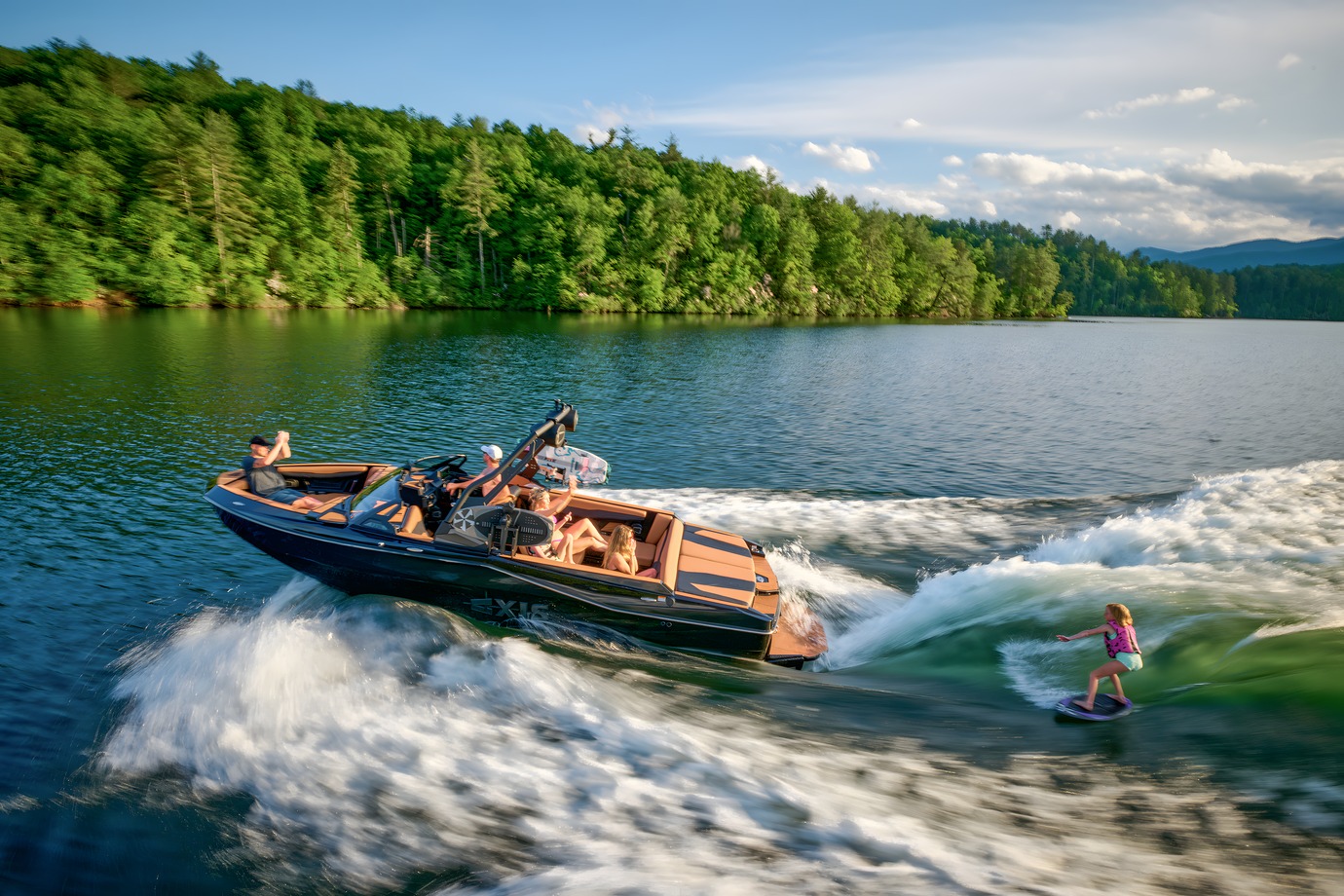 A black Axis T235 with tan interior tows a young wakesurfer across a tree-lined lake under a blue sky.