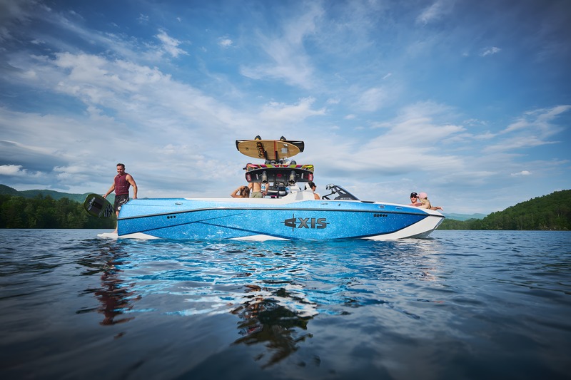 A man stands on the swim platform of a white-and-blue Axis T250 holding a board, wakeboards on the tower, under a big cloudy sky on a calm lake.