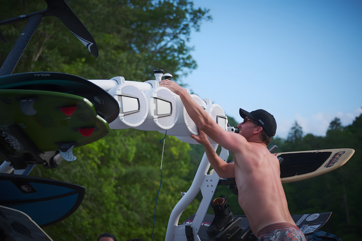 A man in a cap loads a wakeboard into the white tower rack of an Axis T250, boards stowed overhead against a blue sky.