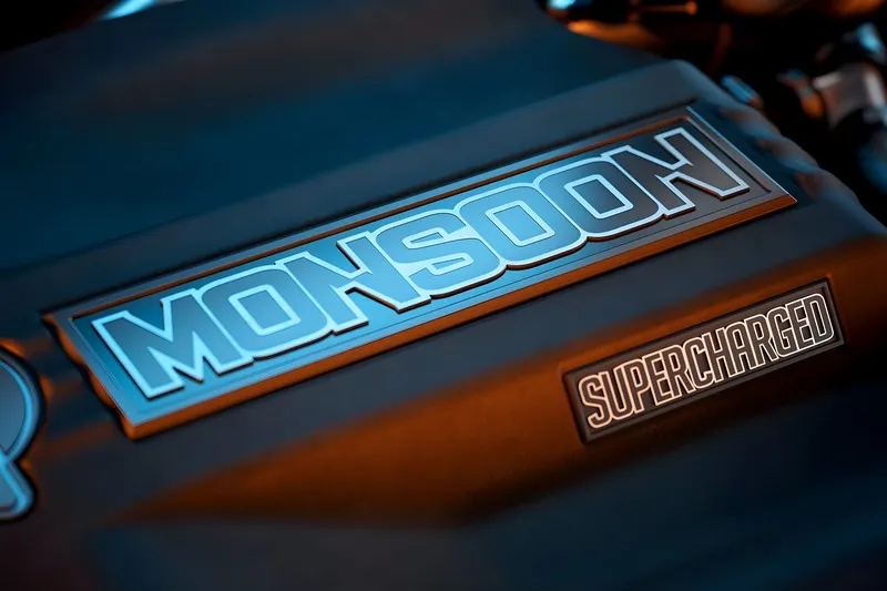 Moody close-up of a black Monsoon engine cover badged 'Monsoon' and 'Supercharged', lit with blue and orange highlights.