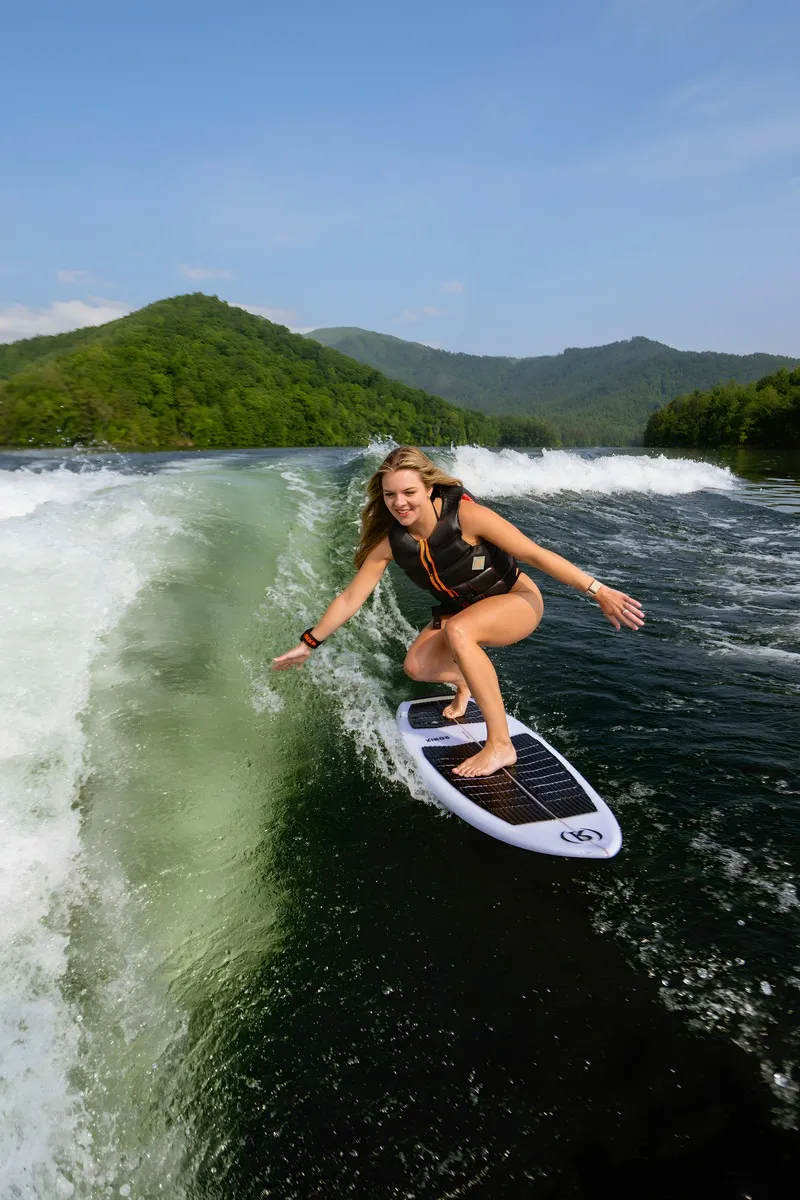 A woman wakesurfs a clean wave on a white board behind an Axis wake boat, forested mountains and blue sky behind.