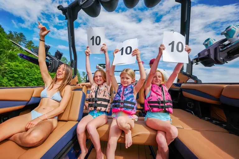A woman and three children in life vests hold up '10' scorecards and cheer from the tan interior of an Axis A245 under a blue sky.