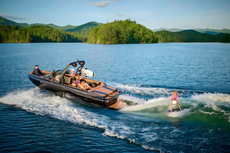 A black Axis T235 runs in side profile across a mountain lake, a young wakesurfer riding its wake under a blue sky.