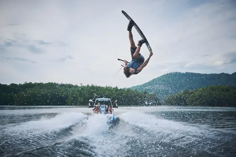 A wakeboarder flips inverted high above the wake behind an Axis wake boat, passengers watching from below on a mountain lake.