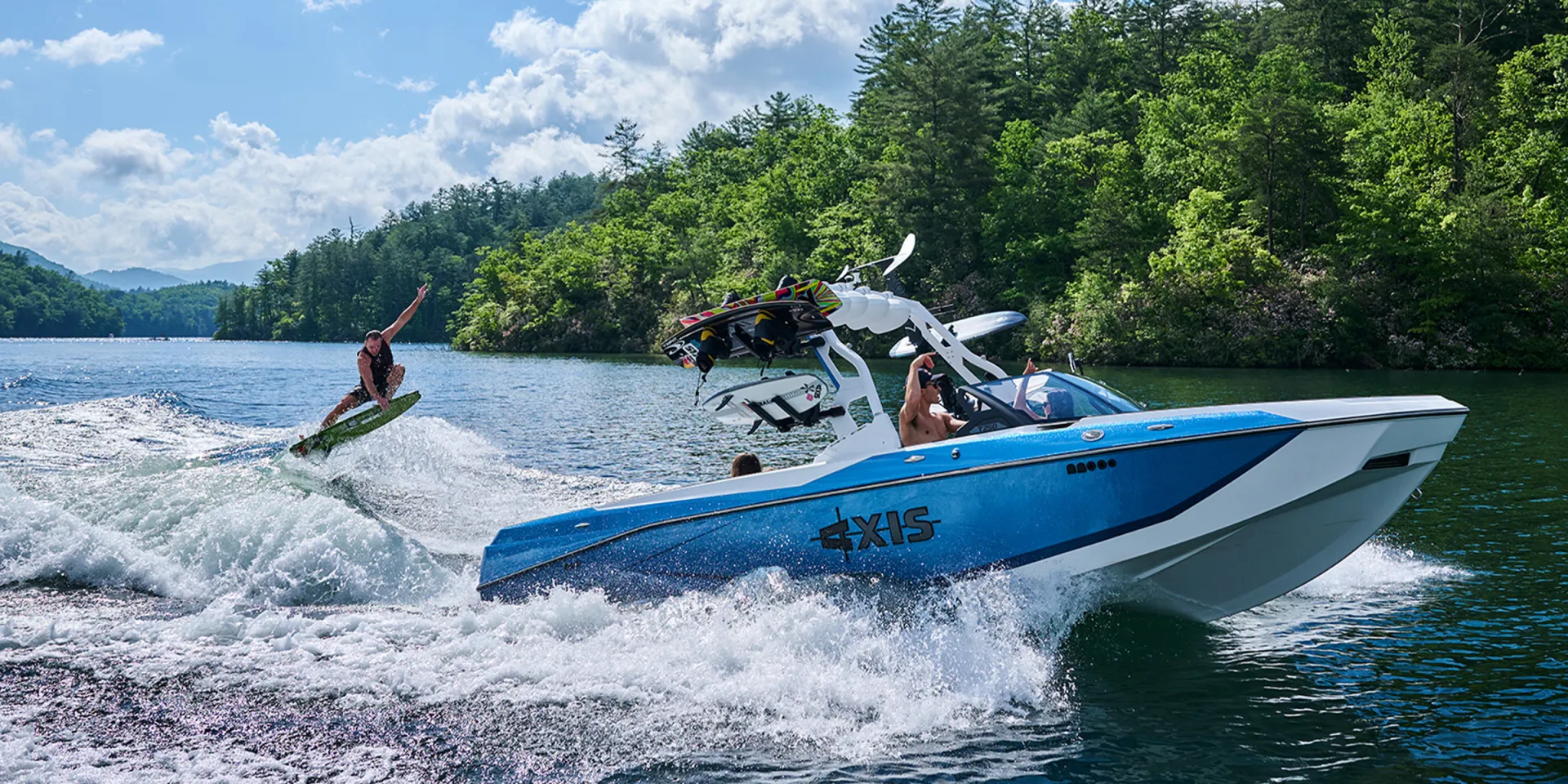 A blue-and-white Axis T250 wake boat carves across a forested mountain lake, towing a wakesurfer who rides the wake with one arm raised while boards sit racked on the tower.