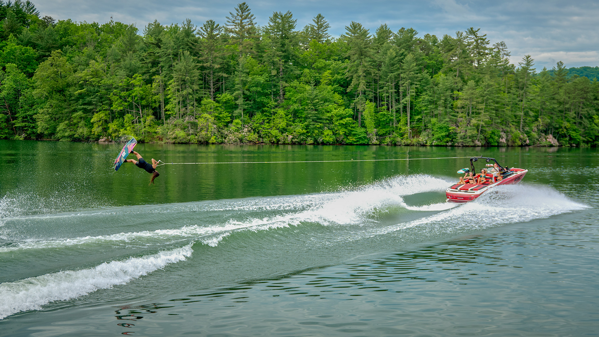 A wakeboarder launches a big inverted trick off the wake behind a red Axis A225, passengers watching from the boat with a wooded shoreline behind.