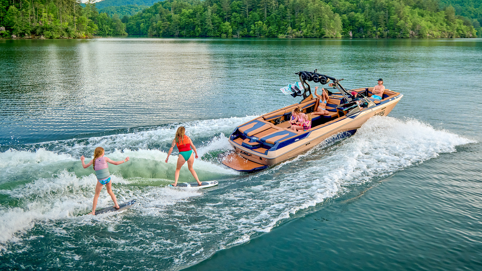 Two wakesurfers ride the wake behind a bronze Axis A245 as passengers cheer from aboard, forested hills and a calm green lake behind them.