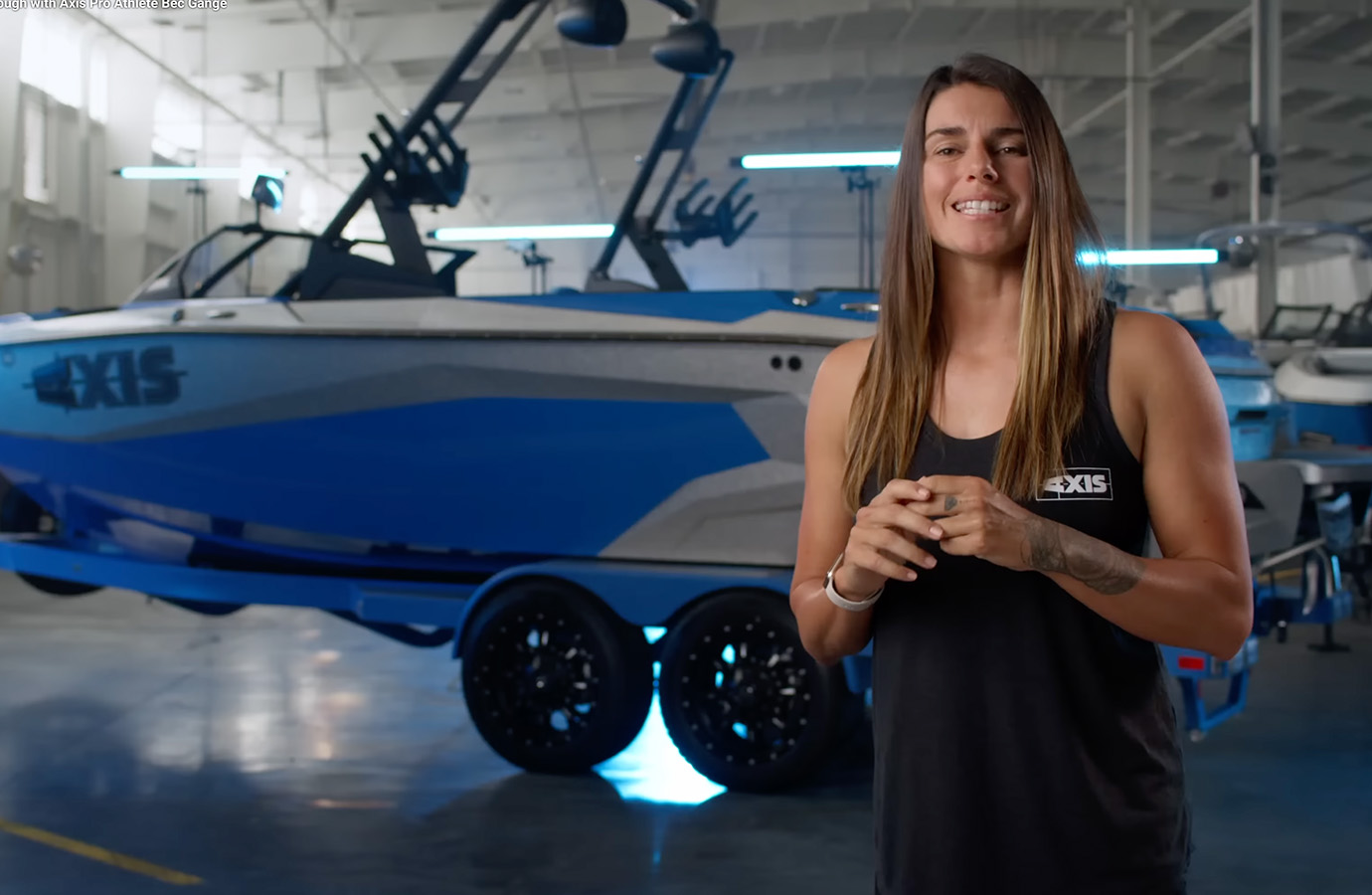 Axis pro athlete Bec Gange speaks to camera in a boat warehouse, a blue and silver Axis A225 on its trailer behind her under bright lights.