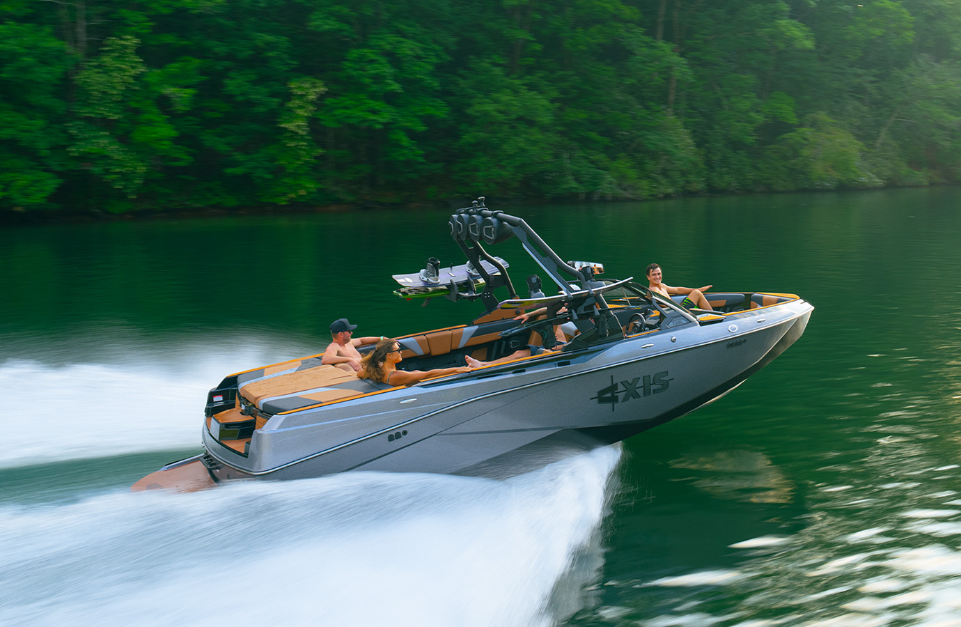 An Axis T235 with a silver hull and tan interior carves across a green lake at speed, several riders aboard and spray kicking up along the forested shore.