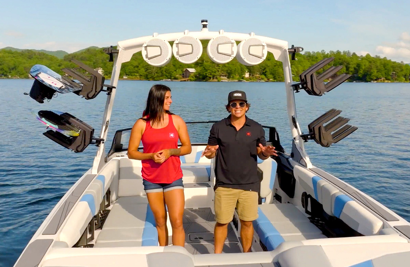 Two walkthrough hosts stand smiling in the white and blue cockpit of an Axis T250, wakeboards on the tower rack and a mountain lake behind them.