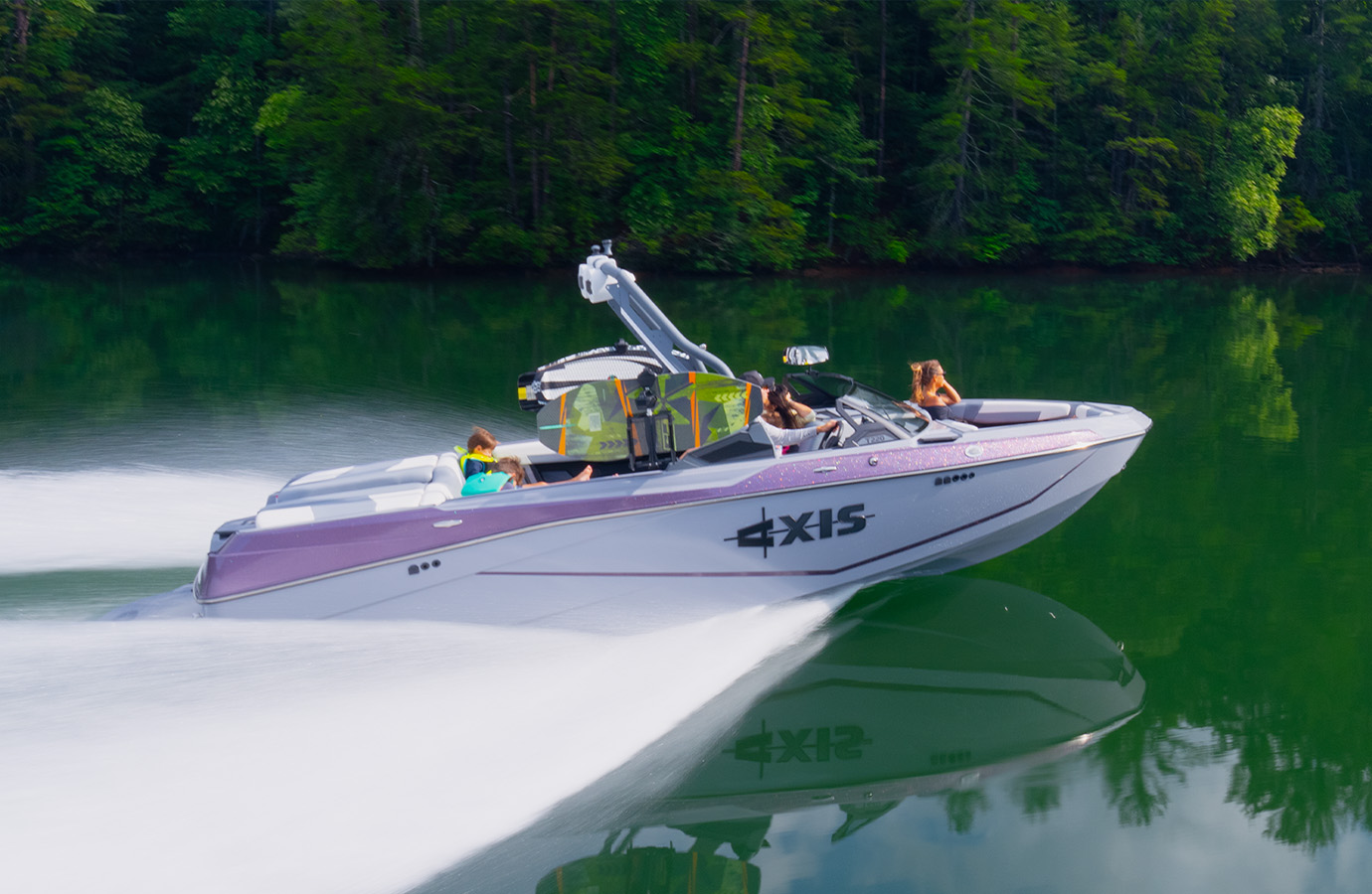 An Axis T220 with a purple and white metalflake hull cruises across a calm green lake, family aboard and a wake trailing behind, forested shoreline behind.