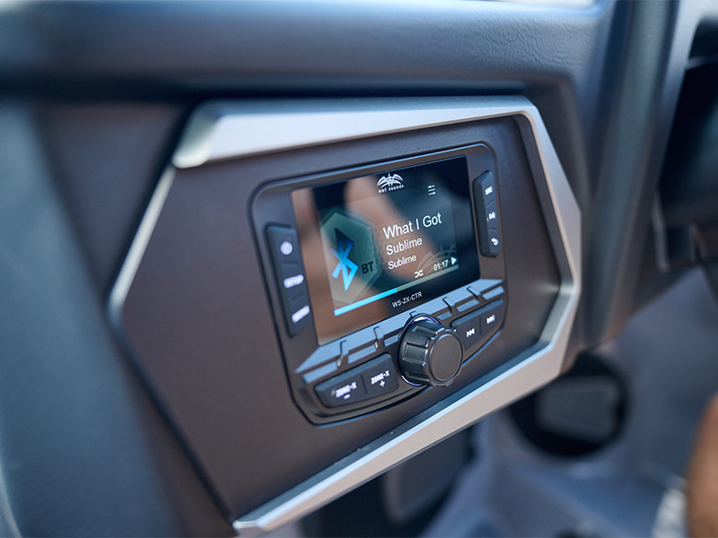 Close-up of the Axis WET Sounds stereo head unit showing a song playing over Bluetooth on its color display, set into the dark dash panel.