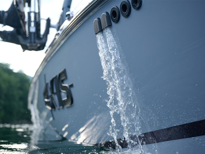 Two streams of water pump out of the ballast drain ports on the side of a gray Axis hull at the waterline, the Axis logo and forested shore behind.