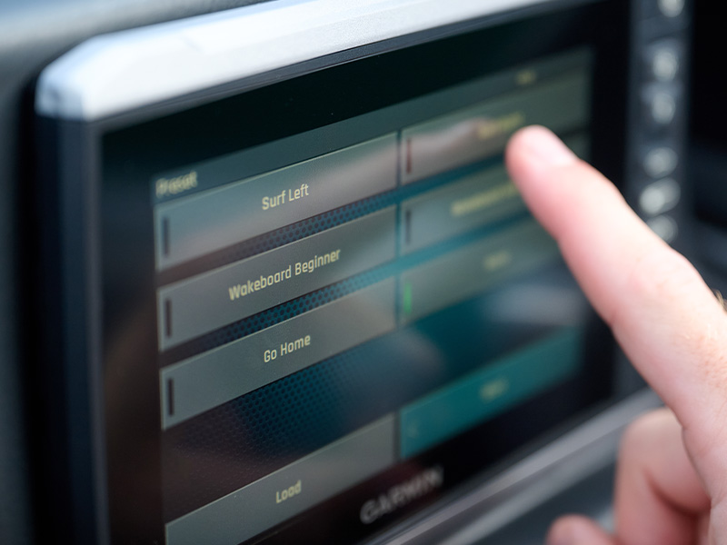 A finger reaches toward the Axis Garmin touchscreen displaying ride presets including Surf Left, Wakeboard Beginner, and Go Home for the boat.