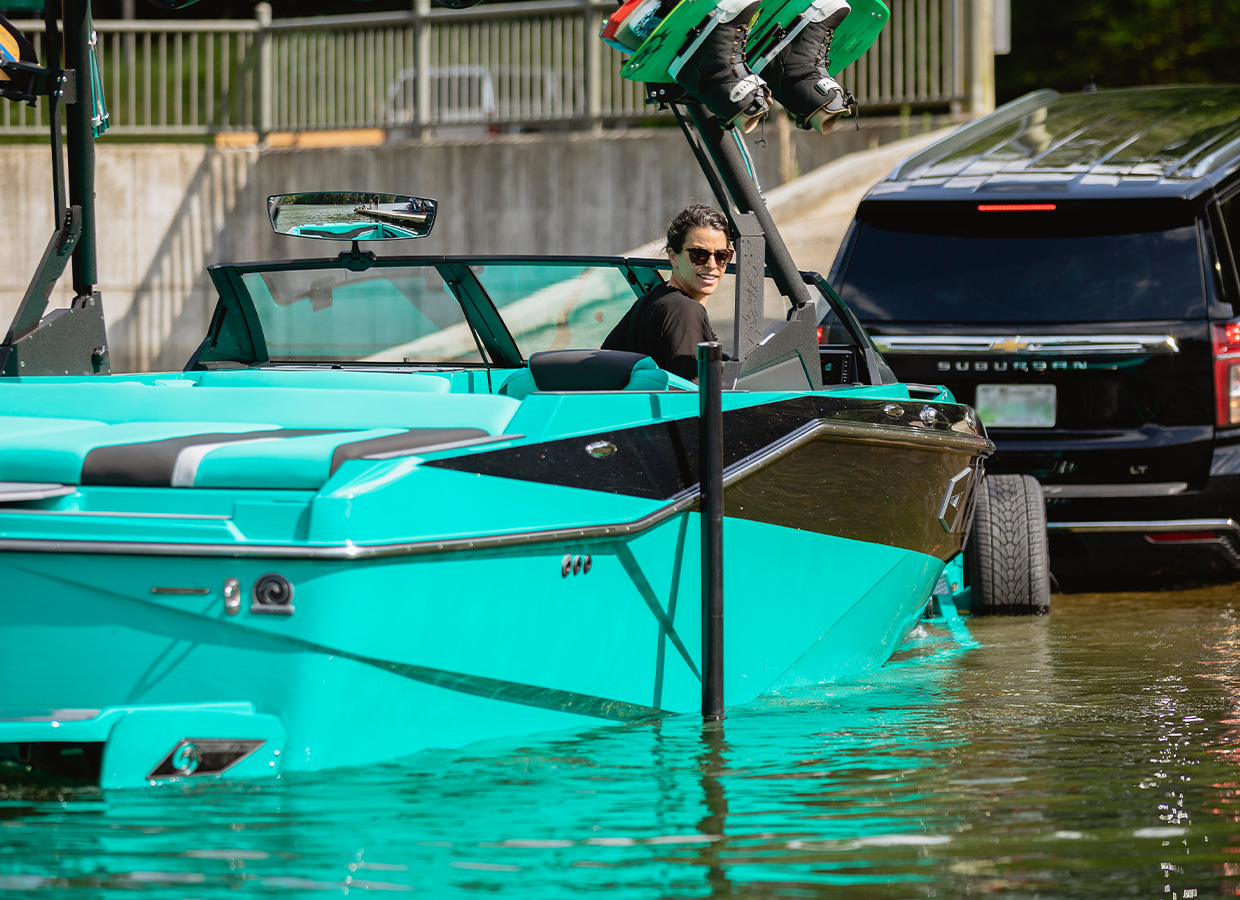 A woman drives in a teal Axis T220 as it loads onto its trailer behind a black SUV at a concrete boat ramp, wakeboards stowed on the tower rack.