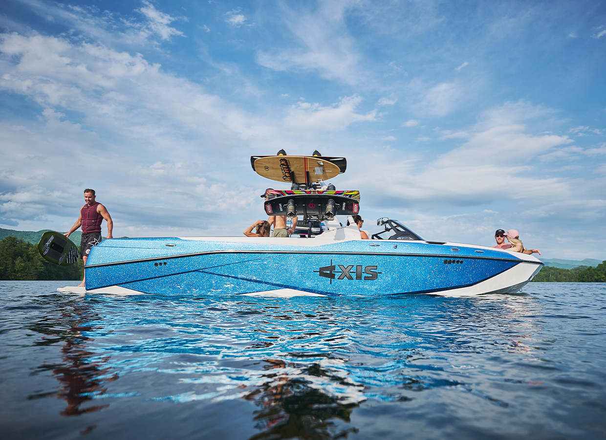 An Axis T250 with a sparkling blue metalflake hull sits on calm water as a rider in a maroon vest steps onto the swim platform with a wakeboard.