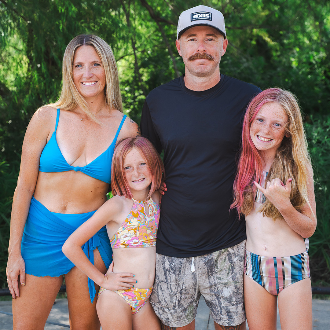 The Neal family, Axis ambassadors, pose together in swimwear in front of leafy green trees — the dad in an Axis cap, the mom in a blue two-piece, and their two young daughters with pink-streaked hair, one throwing a shaka.