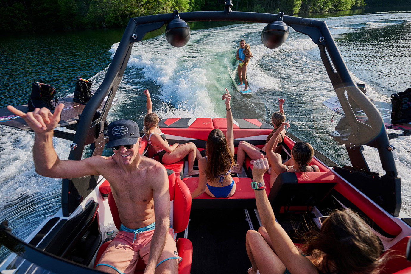 Friends cheer and throw shakas from the red interior of an Axis A-Series, gathered at the sliding Skybox seat as a wakesurfer rides behind on a forested lake.