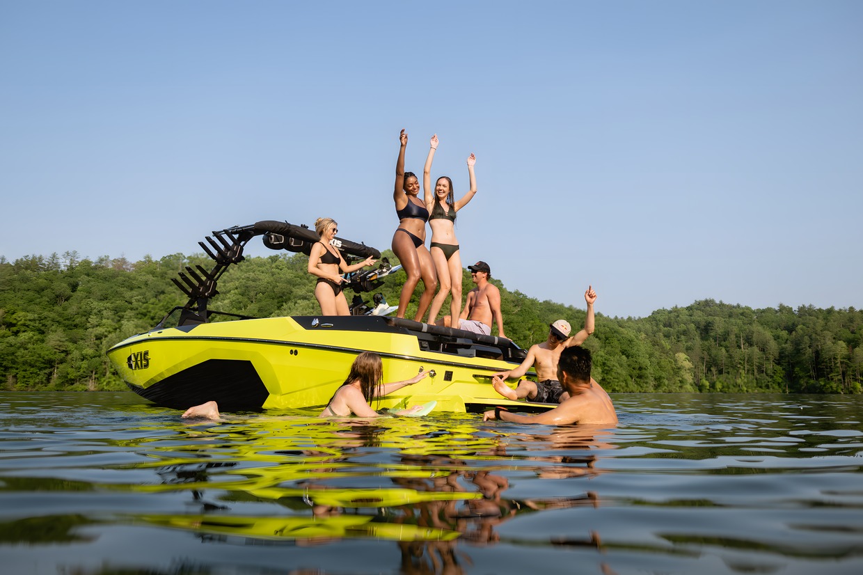 Friends dance and cheer on the transom of a lime-green Axis wake boat near a forested shore, others swimming alongside.
