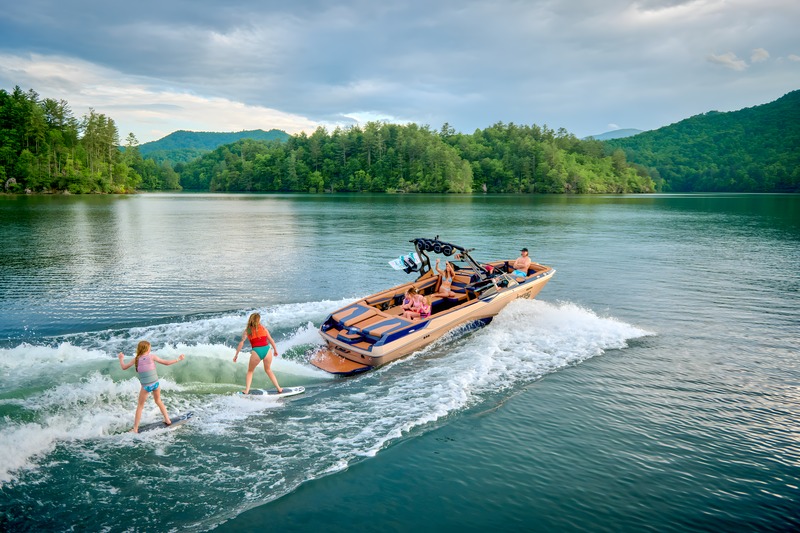 A navy-and-tan Axis A245 tows two young wakesurfers across a calm lake ringed by forested hills.