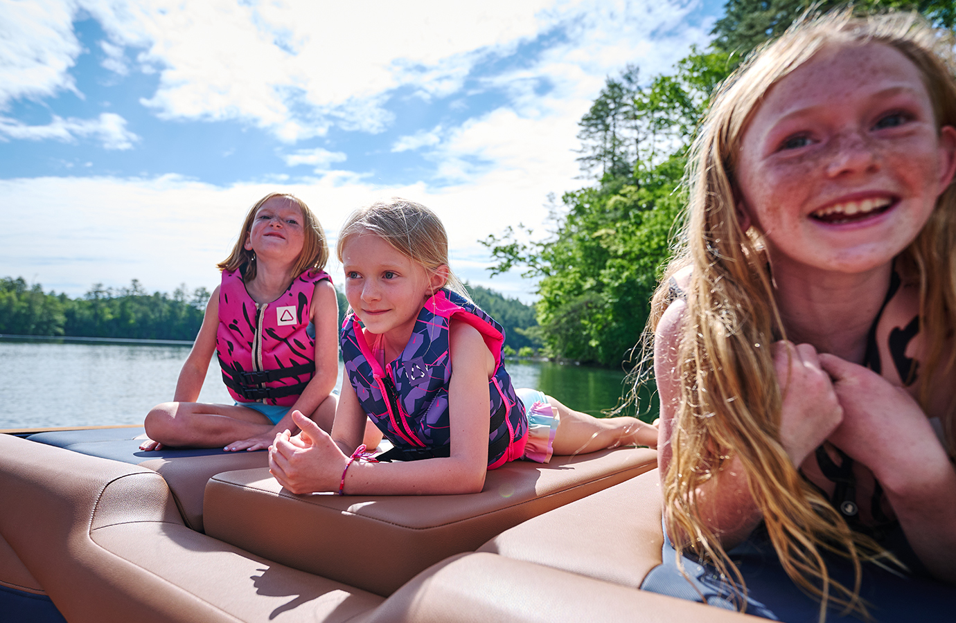 Three girls in life vests relax on the tan bow walkover cushion of an Axis A245, one grinning close to the camera, forested lake behind.