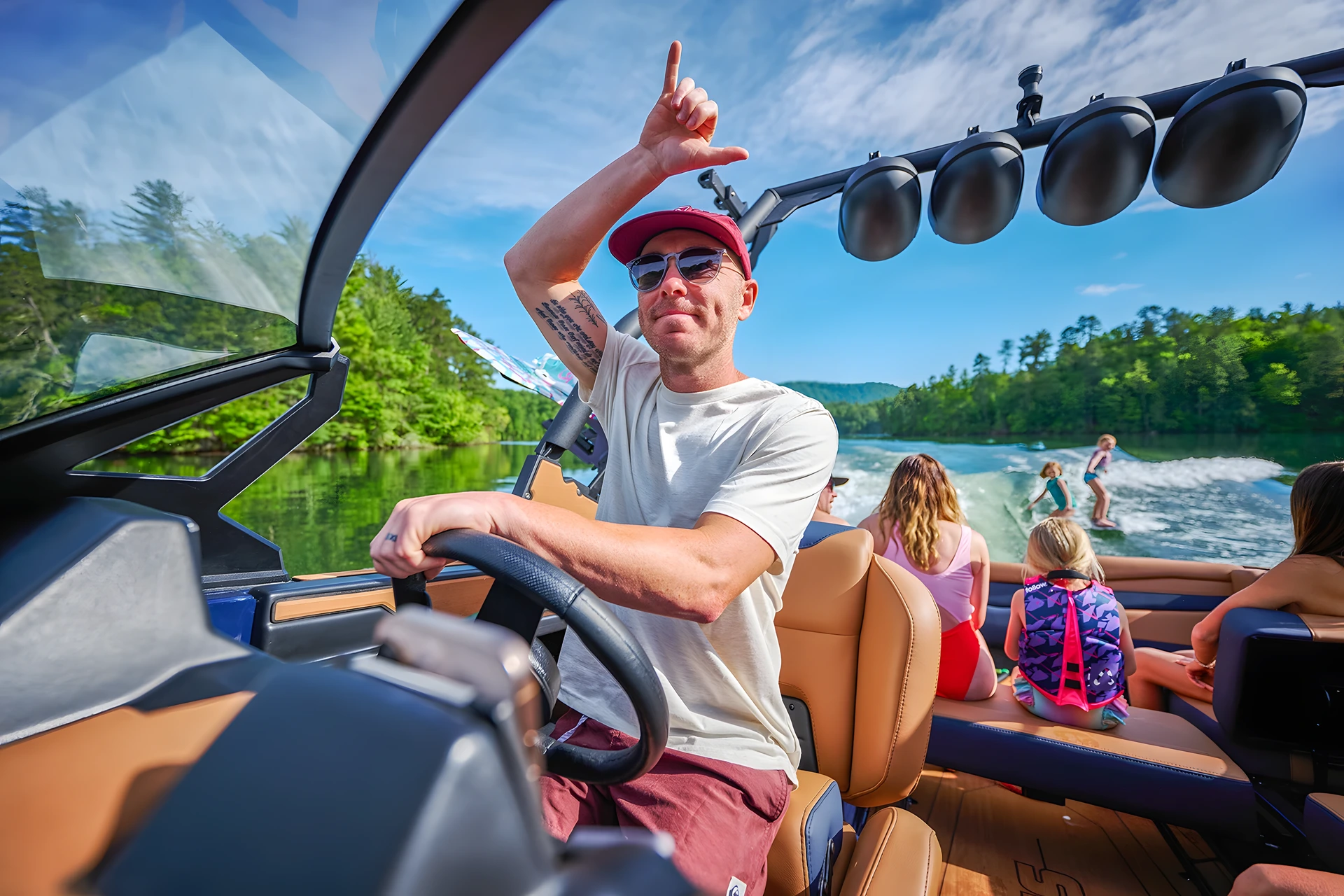 From the bow, a driver at the Axis helm points skyward as passengers watch a wakesurfer behind the boat, tan interior and forested mountains around a calm lake.