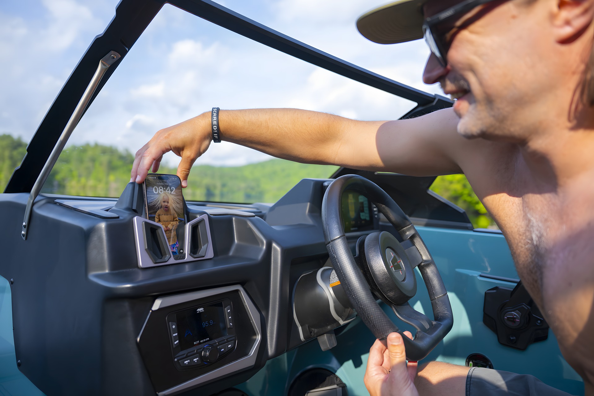 A driver in a cap and sunglasses sets his phone into the wireless charging cradle on the Axis Dash, one hand on the wheel, a forested lake beyond the windshield.