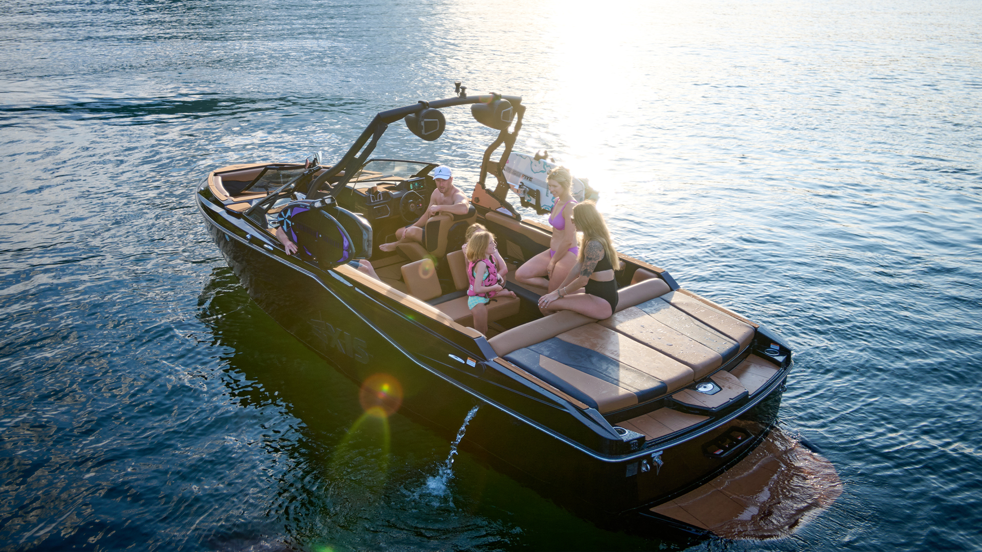 A dark-hulled Axis wake boat idles on a calm lake in warm evening light, friends relaxing aboard as the sun glints off the water.