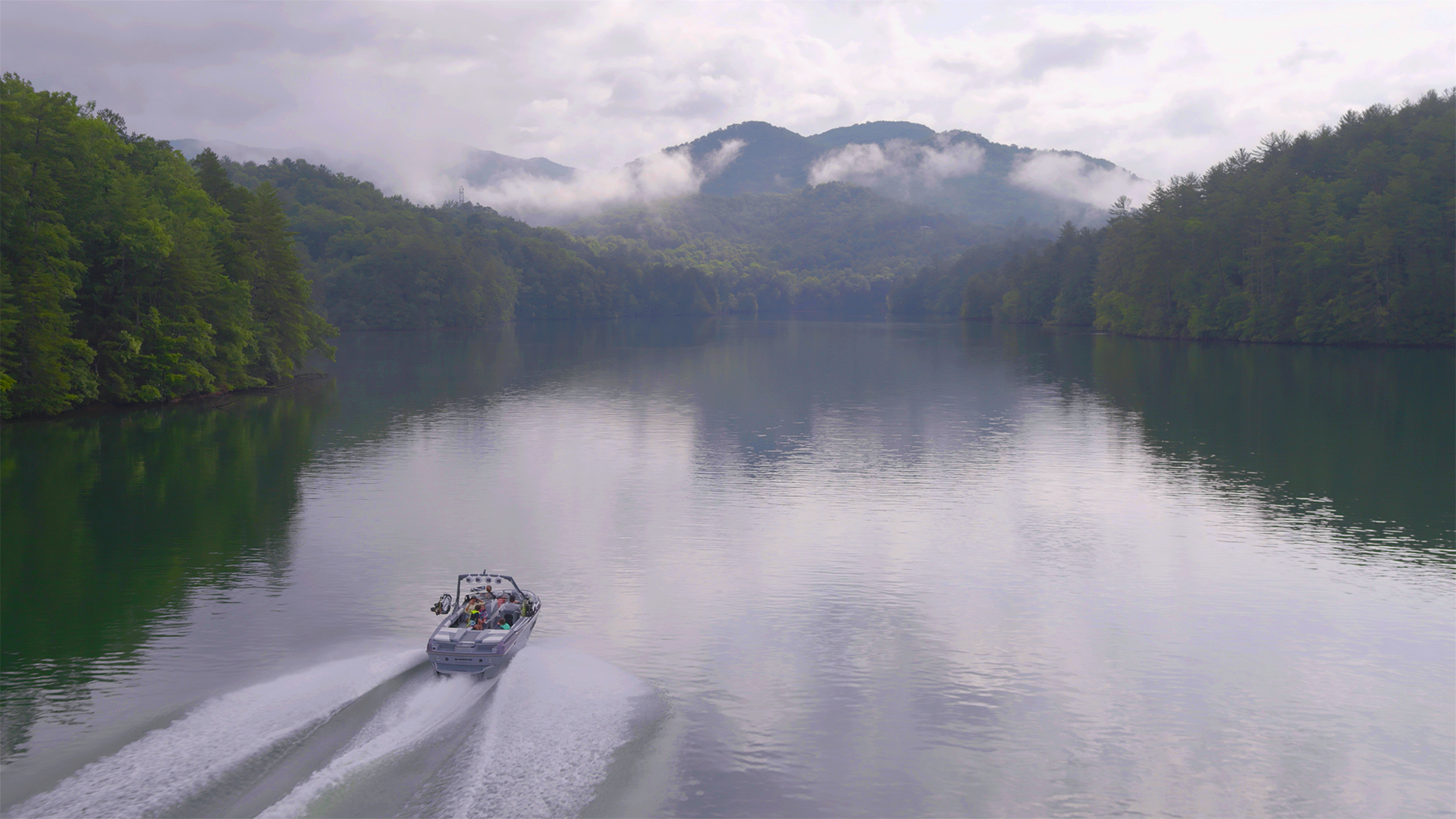 Aerial view of an Axis wake boat cutting across a calm lake below mist-shrouded forested mountains.