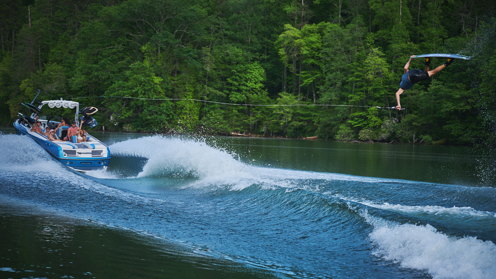 A blue Axis wake boat tows a wakeboarder who launches a big inverted trick off a heavy wake on a forested lake.