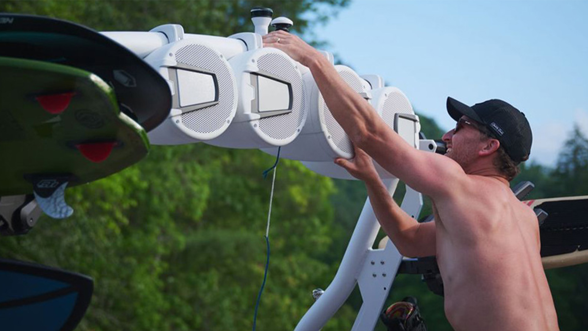 A man in a cap loads a wakeboard into the white tower rack of an Axis T250, boards stowed overhead against a blue sky.