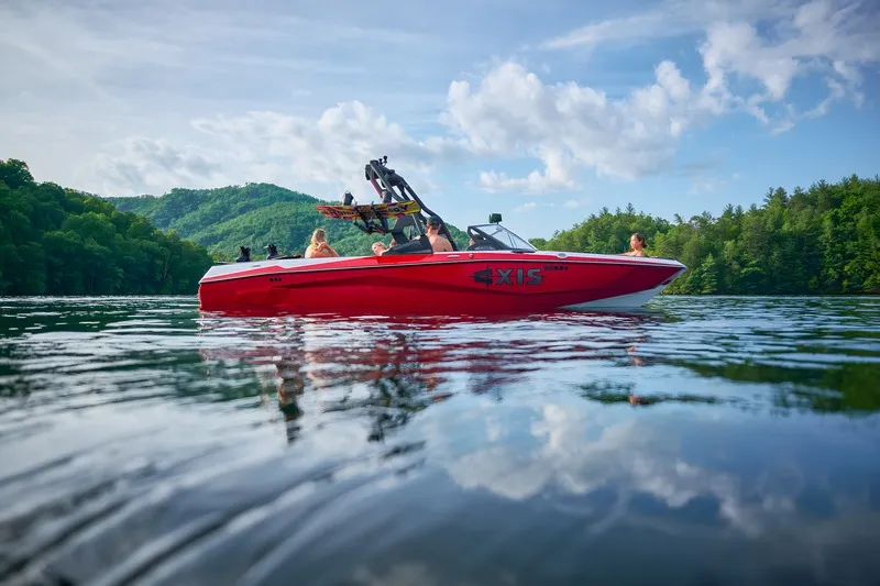 A red Axis A225 idles on a calm lake under a bright cloudy sky, passengers aboard and forested hills reflected in the water.