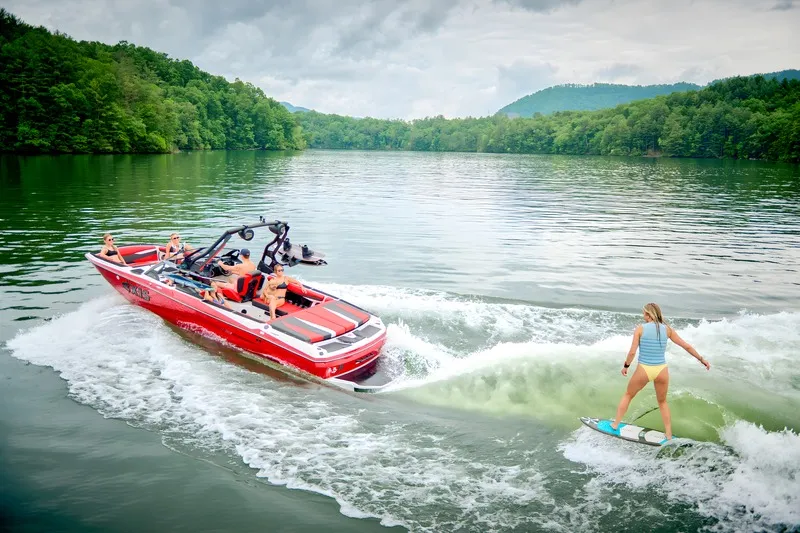 A woman wakesurfs behind a red Axis A225 on a mountain-ringed lake, the boat running alongside her clean wake.
