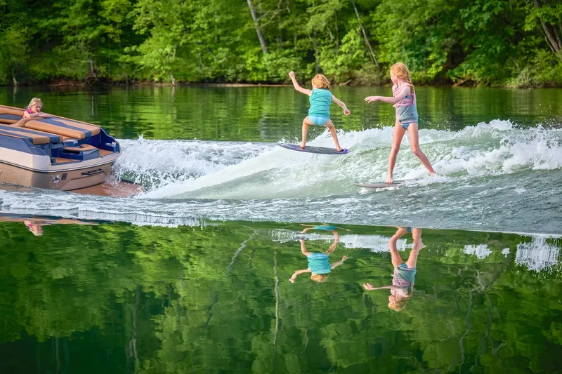 Two children wakesurf side by side on the clean wake behind a navy-and-tan Axis A245, forest mirrored in the glassy water.
