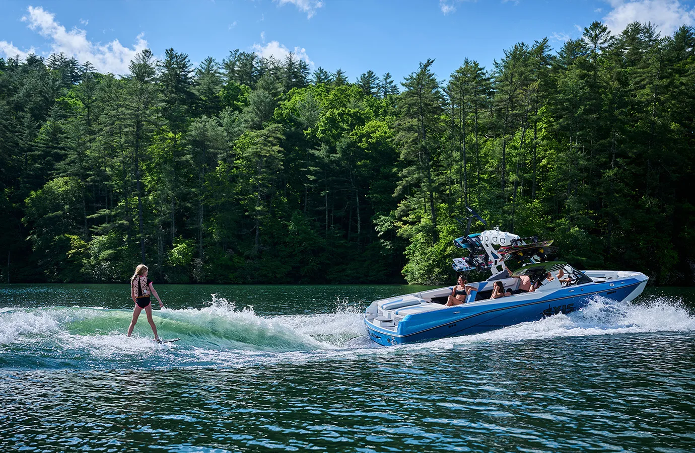 A wakesurfer in a black vest carves a clean green wave behind an Axis T250 on a forested lake.