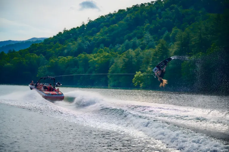 A red Axis A225 tows a wakeboarder who launches a big trick off the wake, forested mountains behind.