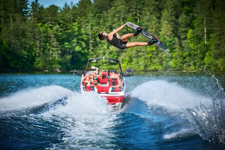 A wakeboarder flips high off the wake behind a red Axis A225, spray bursting on a forested lake.