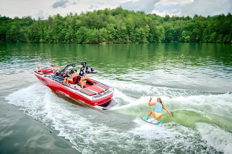 A woman wakesurfs the clean wake behind a red Axis A225 on a calm, tree-lined lake.